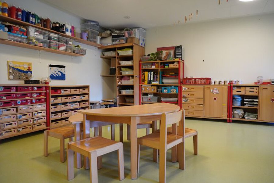 Kreativraum im Kindergarten mit Kindertischen und -stühlen, Regalen voller Bastelmaterialien, Farben und Arbeitsutensilien.