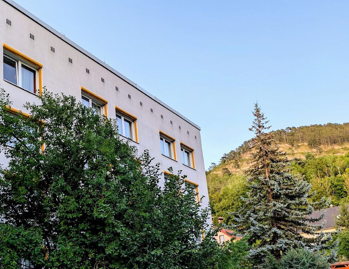 Modernisierte Außenansicht des Akademiehotels mit heller Fassade, gelben Fensterrahmen und umgebender Begrünung vor bewaldetem Hang