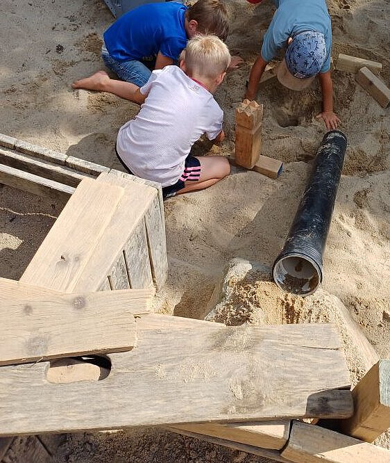 Kinder spielen im Sandkasten mit Holzstücken und Rohren.