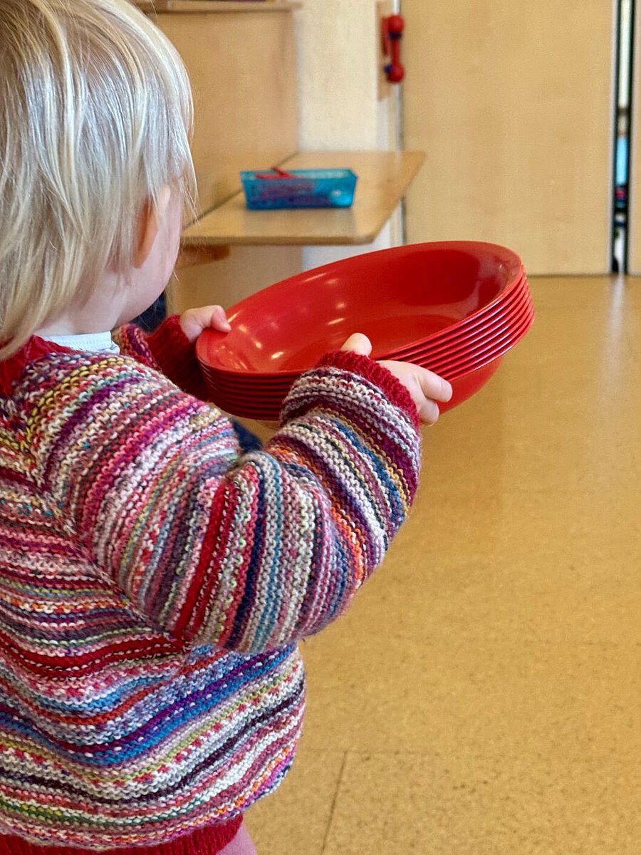 Ein Kind trägt mehrere Teller durch den Gruppenraum und übernimmt eine Aufgabe beim Tischdecken. Alltagsbeteiligung stärkt Verantwortungsbewusstsein und Selbstwirksamkeit.
