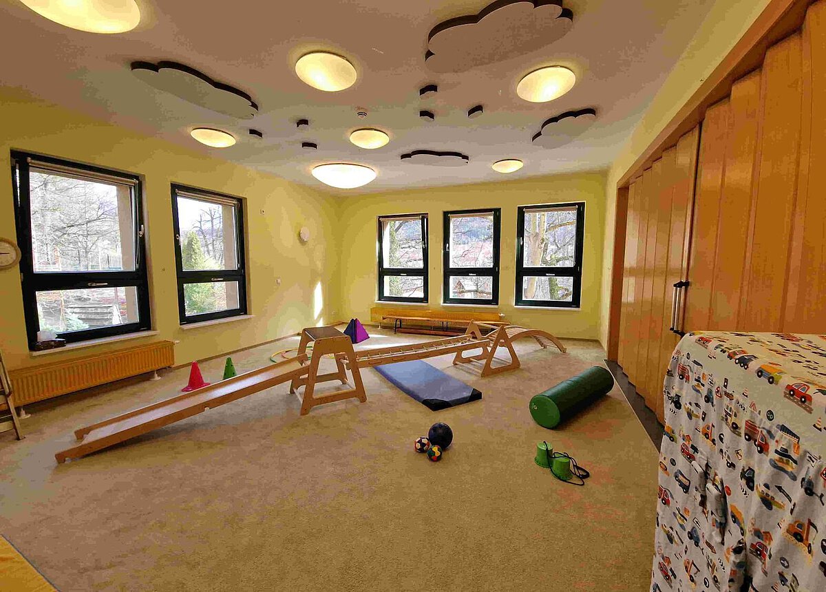 Heller Bewegungsraum im Kindergarten Burgweg mit großen Fenstern, Holz-Sprossenwänden und verschiedenen Spiel- und Motorikelementen wie Balancierbrettern, Turnbänken, Matten, Kegeln und einer grünen Rolle auf Teppichboden.