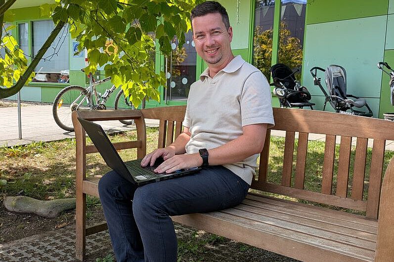 Arbeiten im Grünen auf dem Gelände der Kindertagesstätte Grunaer Kinderwelt Interviewpartner Martin Garten mit Laptop auf einer Holzbank unter einem Baum vor dem modernen Kita-Gebäude der Grunaer Kinderwelt mit grüner Fassade