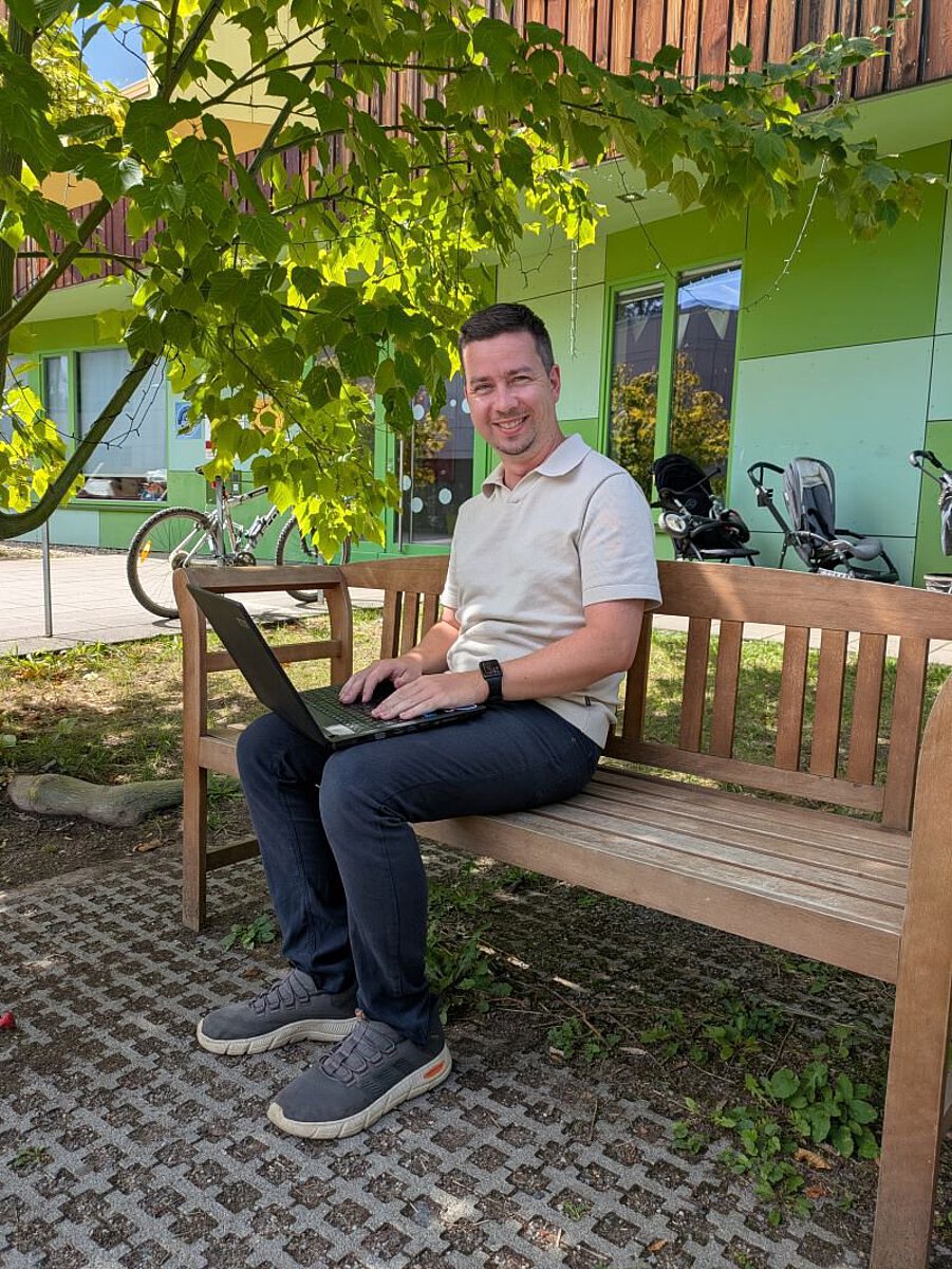 Interviewpartner Martin Garten mit Laptop auf einer Holzbank unter einem Baum vor dem modernen Kita-Gebäude der Grunaer Kinderwelt mit grüner Fassade