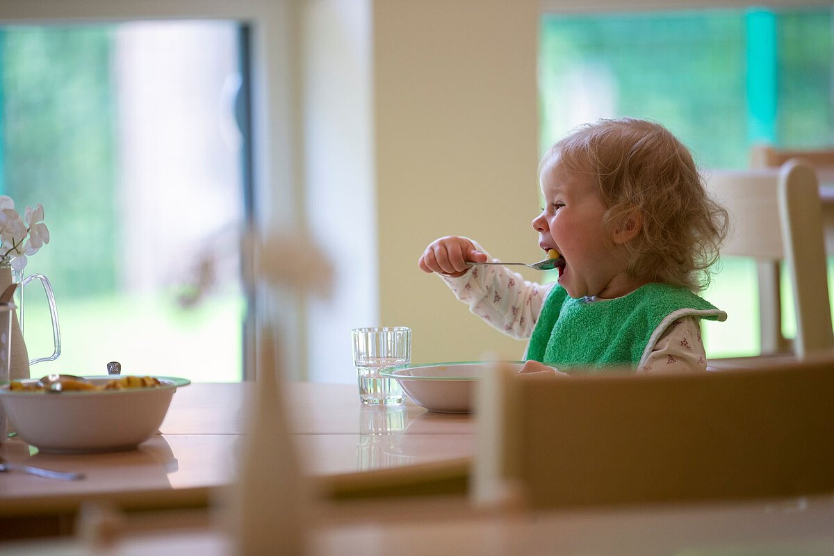 Tisch in einem hellen Raum. Auf dem Tisch stehen eine Schüssel mit Essen, ein Glas Wasser und eine Karaffe. Ein Kind sitzt auf einem Stuhl und hält einen Löffel über einer Schüssel. Im Hintergrund sind große Fenster, durch die grünes Gras zu sehen ist
