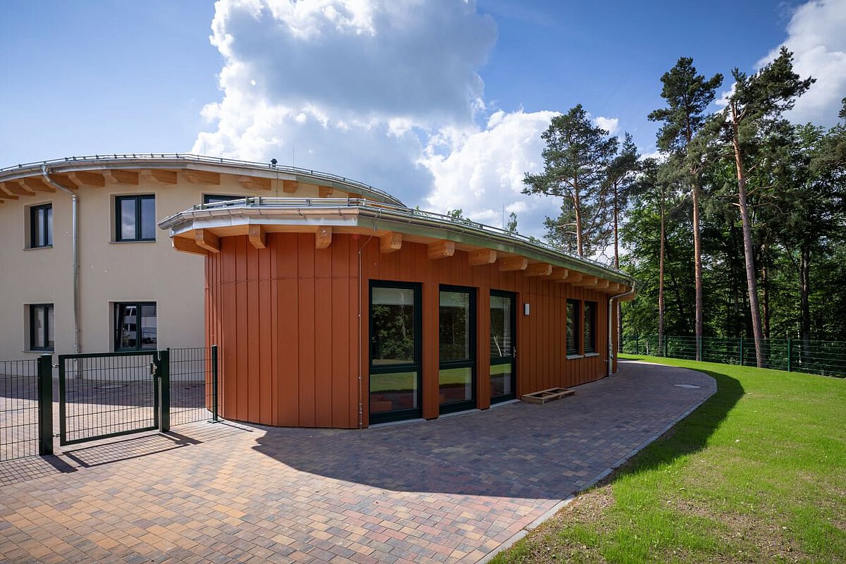 Das Bild zeigt ein modernes Gebäude mit runden Formen. Die Fassade ist orange und hat große Fenster. Vor dem Gebäude liegt ein gepflasterter Weg. Rechts ist eine grüne Wiese und dahinter stehen hohe Bäume. Der Himmel ist blau mit weißen Wolken.