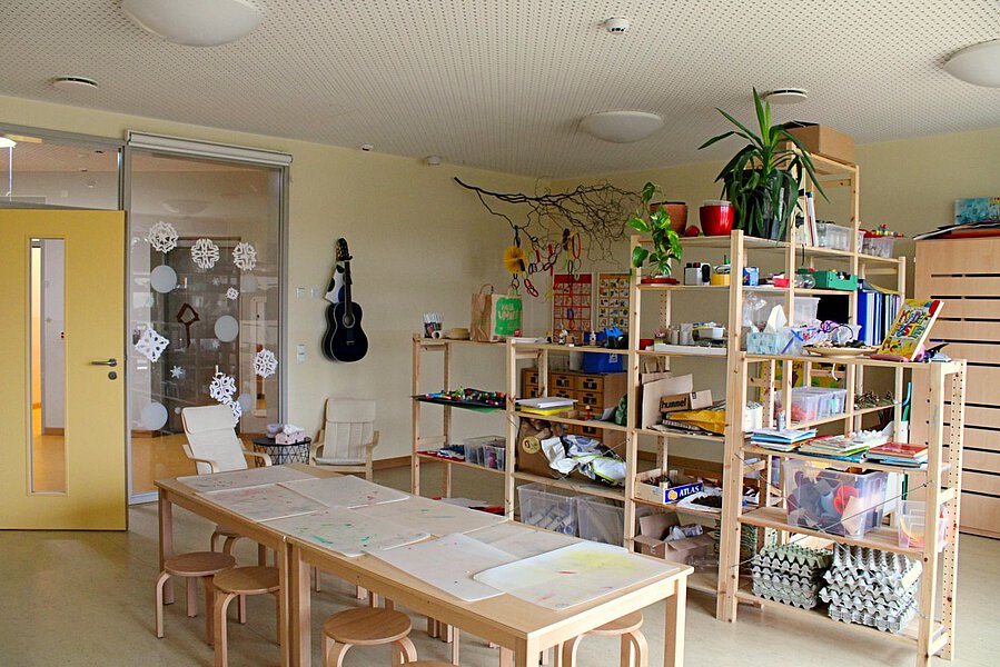 Zu sehen ist ein im Raum freistehender großer Tisch mit Stühlen. Im Hintergrund steht ein Regal mit verschiedenen Materialien zur Selbstentnahme für die Kinder zum kreativen Gestalten.