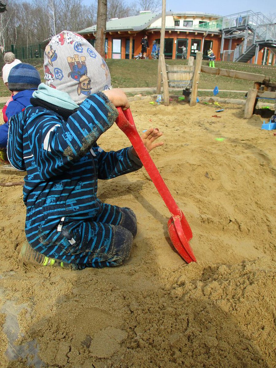  Ein Kind sitzt im Sandkasten. Es trägt einen blau gestreiften Anzug und eine bunte Mütze. Das Kind hält eine große rote Schaufel und gräbt im Sand. Im Hintergrund spielen andere Kinder. Dort steht auch ein Gebäude mit orange Wänden und grünem Dach. Es ist draußen, der Himmel ist grau.