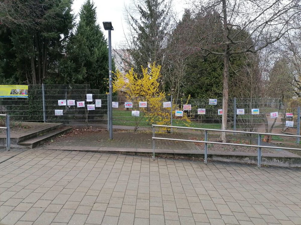 Mehrere bunte Flaggen und Schilder mit verschiedenen Sprachen hängen an einem Bauzaun vor dem Kindergarten, um kulturelle Vielfalt zu zeigen.