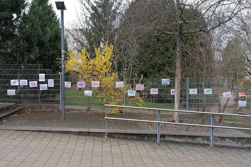 Mehrere bunte Flaggen und Schilder mit verschiedenen Sprachen hängen an einem Bauzaun vor dem Kindergarten, um kulturelle Vielfalt zu zeigen.