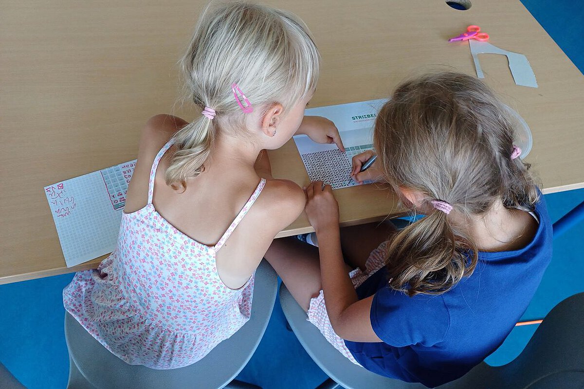 Zwei Kinder sitzen an einem Tisch und arbeiten gemeinsam an einem Blatt mit Kästchenmuster.