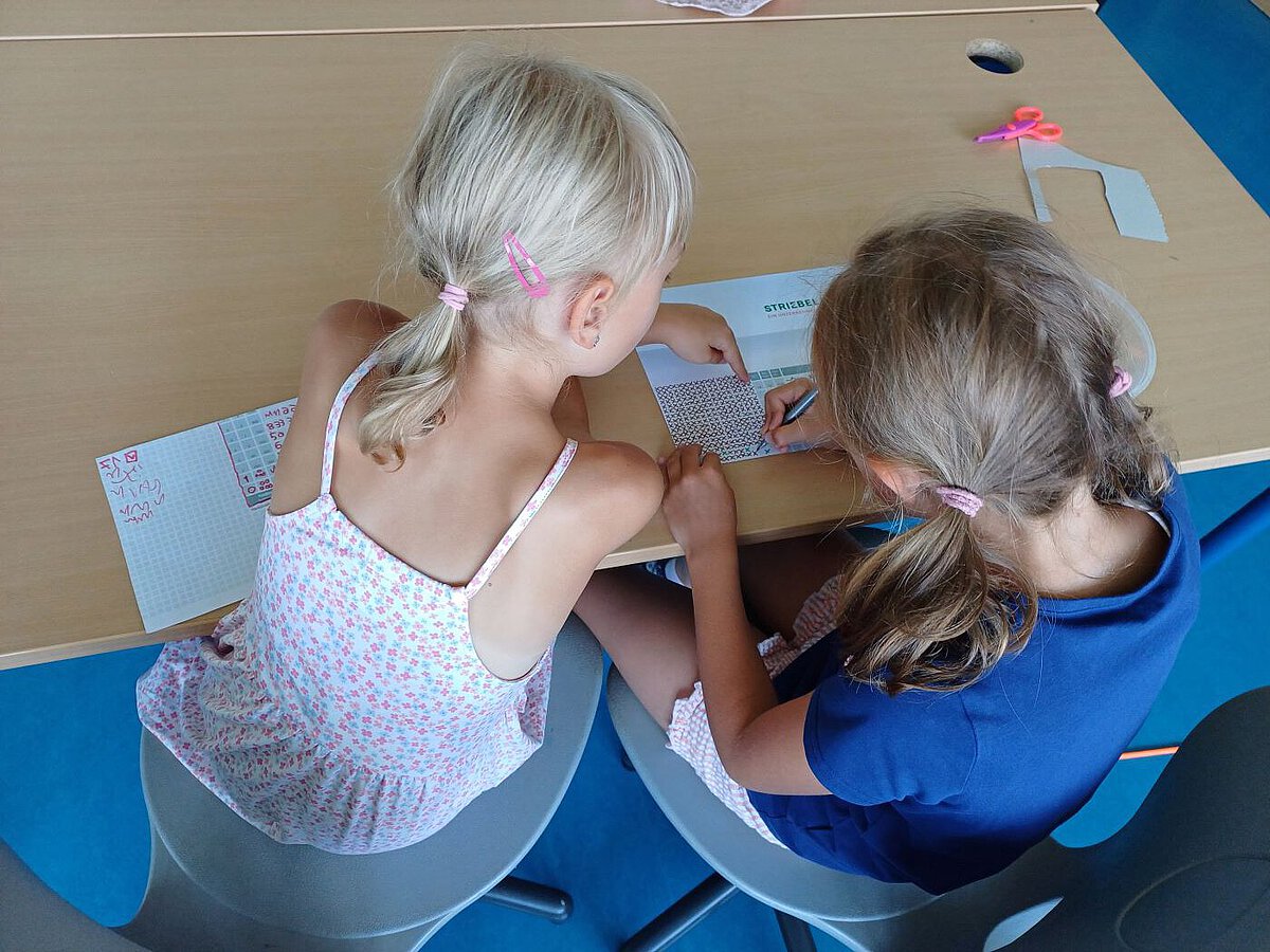 Zwei Kinder sitzen an einem Tisch und arbeiten gemeinsam an einem Blatt mit Kästchenmuster.