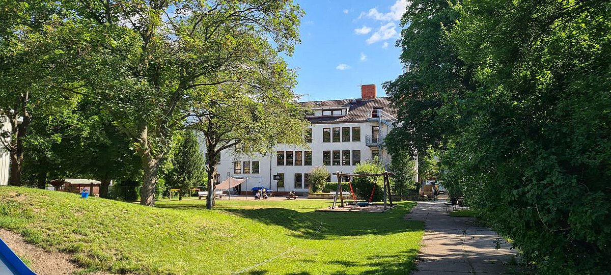 Blick auf das Außengelände des Kindergartens Vollbrachtfinken mit Grünflächen, Bäumen, Spielgeräten und dem Hauptgebäude im Hintergrund.