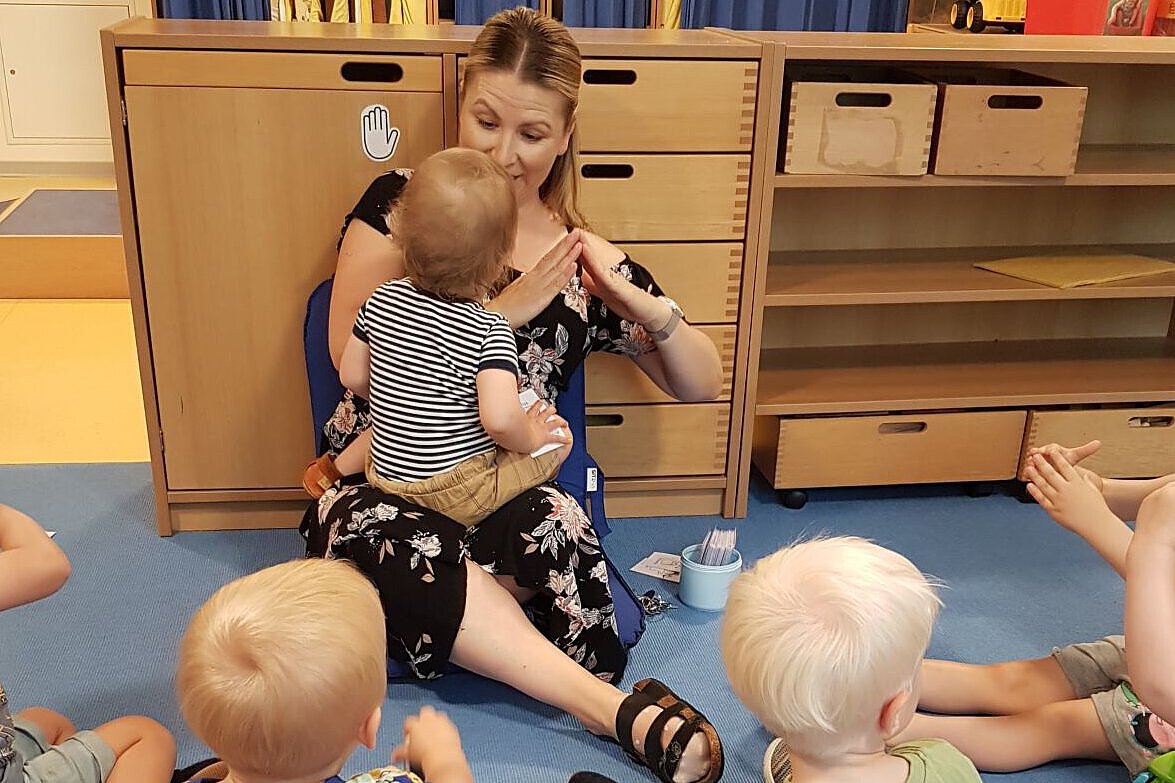 Person sitzt mit mehreren Kleinkindern im Kreis auf dem Boden und begleitet ein Kind durch Gesten und Interaktion.