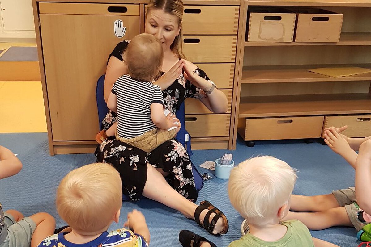 Pädagogische Fachkraft der Kita Hölderlinstraße mit Kleinkindern Person sitzt mit mehreren Kleinkindern im Kreis auf dem Boden und begleitet ein Kind durch Gesten und Interaktion.