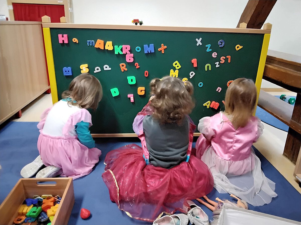Buchstaben - Sprache - Mathematik - Kognitive Entwicklung - Kindergarten Windischholzhausen Drei Kinder sitzen verkleidet als Prinzessinnen vor einer Tafel und betrachten die Buchstaben- und Zahlenmagnete