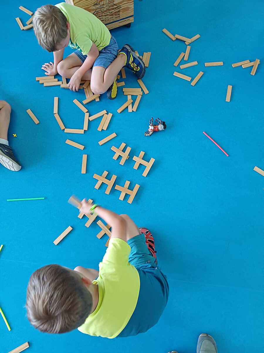 Zwei Kinder sitzen auf blauem Teppich und bauen mit Holzbausteinen geometrische Formen.
