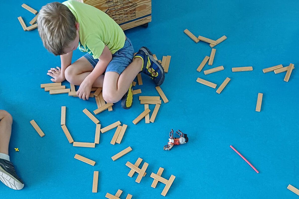 Zwei Kinder sitzen auf blauem Teppich und bauen mit Holzbausteinen geometrische Formen.
