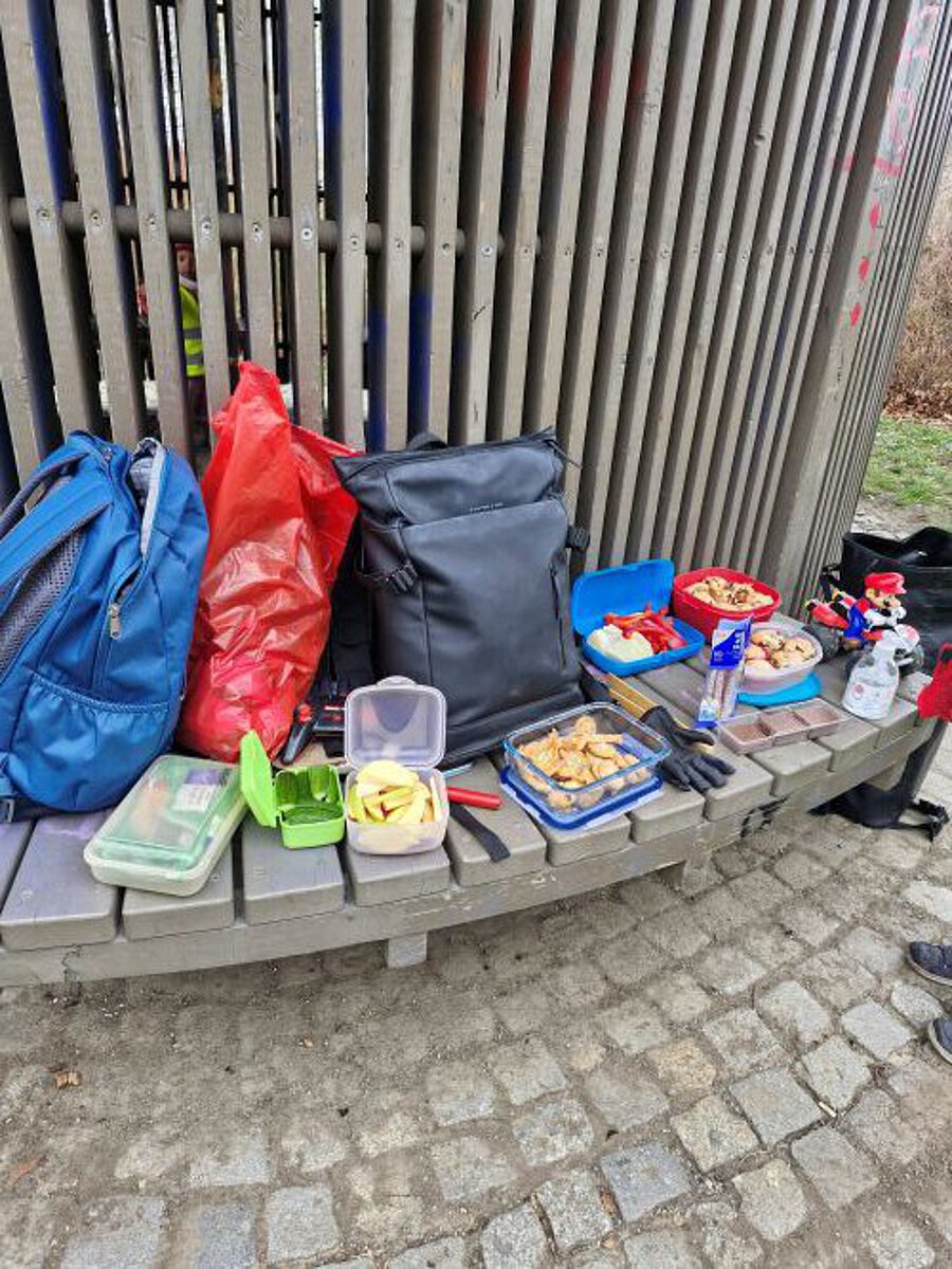 „Mehrere Rucksäcke, ein roter Müllsack und verschiedene Snack‑Dosen stehen auf einer gebogenen Holzbank; davor liegen Lunchboxen mit Obst, Gebäck und kleinen Snacks.“