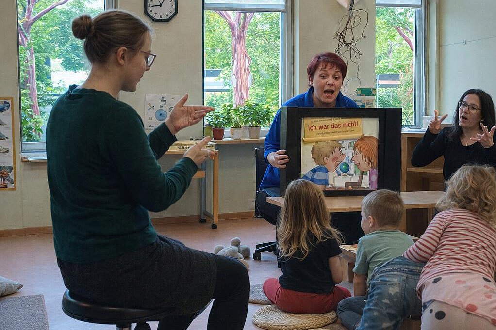 Pädagogische Fachkräfte lesen Kindern in einer Kita eine Geschichte vor und unterstützen diese mit Gebärden; mehrere Kinder sitzen im Halbkreis und verfolgen die Bilder.