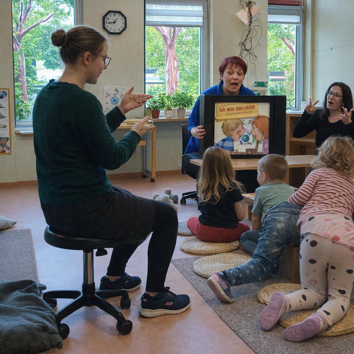 Pädagogische Fachkräfte lesen Kindern in einer Kita eine Geschichte vor und unterstützen diese mit Gebärden; mehrere Kinder sitzen im Halbkreis und verfolgen die Bilder.