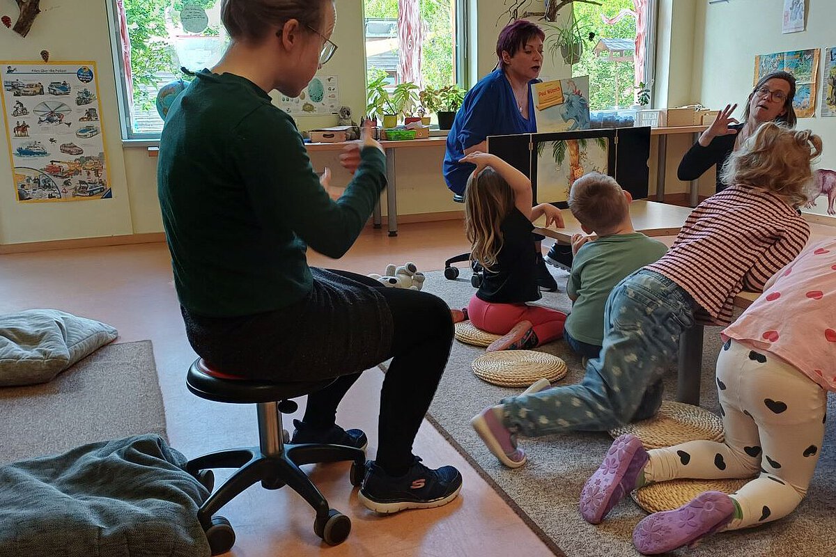 Pädagogische Fachkräfte lesen Kindern in einer Kita eine Geschichte vor und unterstützen diese mit Gebärden; mehrere Kinder sitzen im Halbkreis und verfolgen die Bilder.