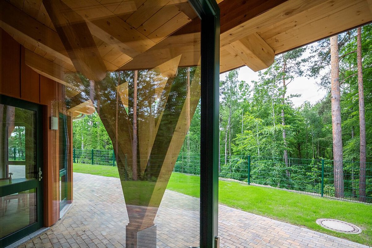 Blick aus dem Kindergarten Am Wald- Klinikum Gebäude in Richtung Wald. Im Vordergrund sind große Glasfenster und ein Holzpfeiler, der das Dach stützt. Draußen liegt ein gepflasterter Weg und eine grüne Wiese. Dahinter stehen viele hohe Bäume mit grünen Blättern. Der Bereich ist von einem grünen Zaun umgeben