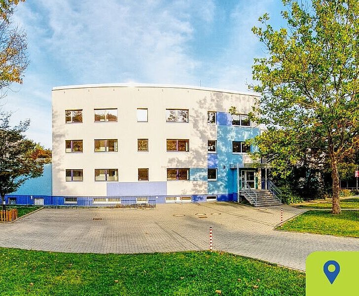 Integrativer Kneipp-Kindergarten Winzerla mit großem Außengelände, Google Maps Verlinkung Modernes, weiß-blaues Kita-Gebäude mit mehreren Etagen, umgeben von grüner Wiese und Bäumen. Im Vordergrund ein gepflasterter Bereich, dahinter Spiel- und Gartenflächen