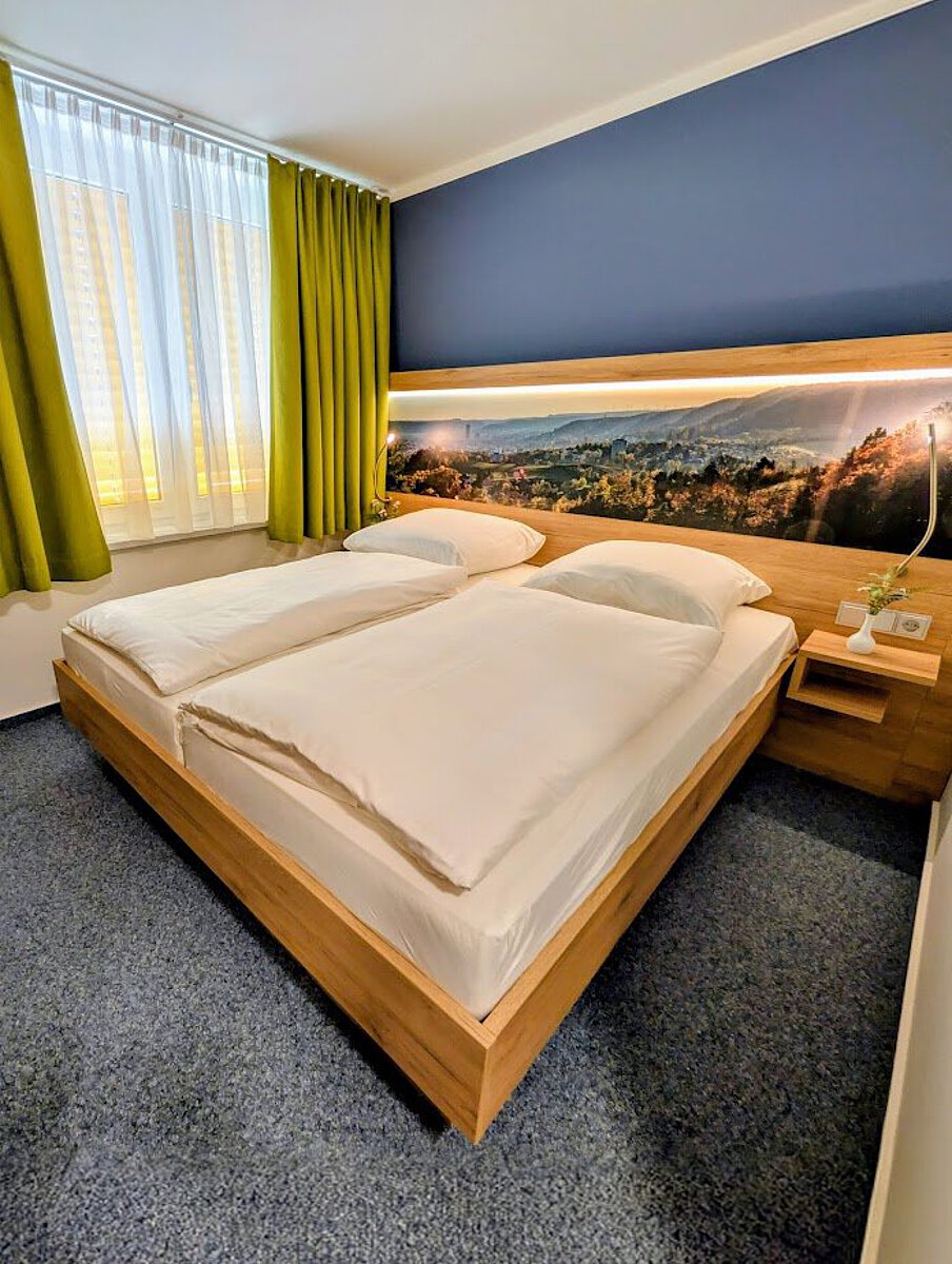 Modern eingerichtetes Doppelzimmer im Akademiehotel mit großem Bett, Holzrahmen, Wandbild mit Landschaftspanorama und Fenster mit grünen Vorhängen