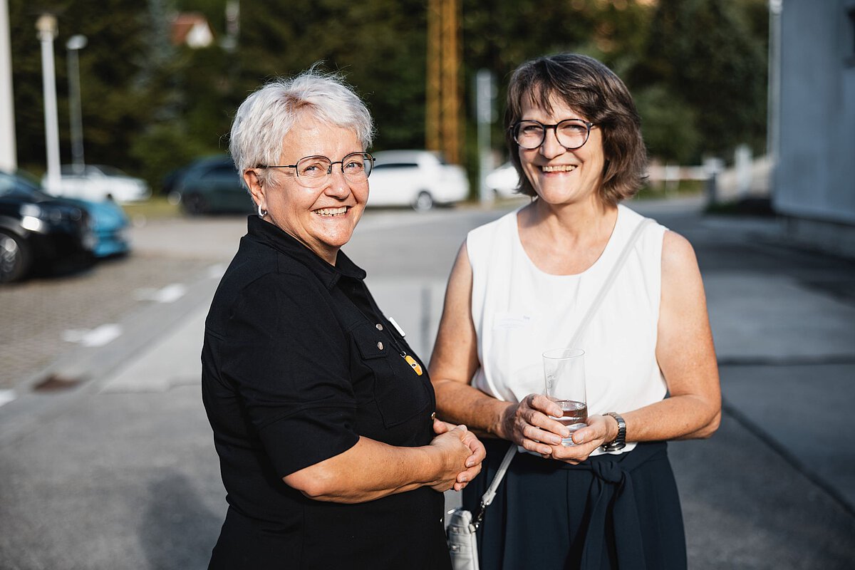Gäste im Gespräch bei Jubiläumsveranstaltung im Freien Zwei Personen stehen im Freien, unterhalten sich und halten Getränke in der Hand während einer Veranstaltung.