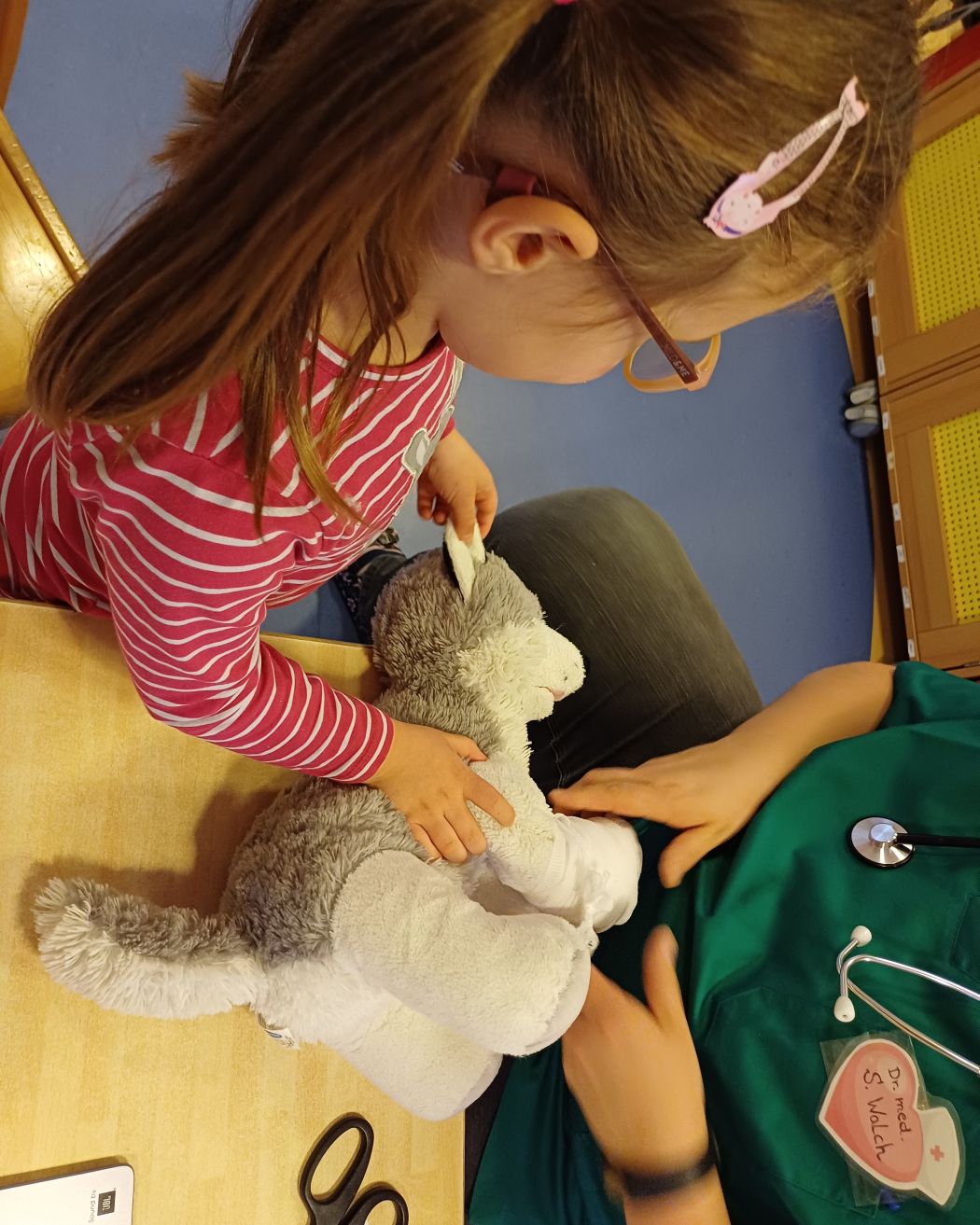 Kind untersucht gemeinsam mit einer erwachsenen Person in grüner Arztkleidung einen Stoffhund