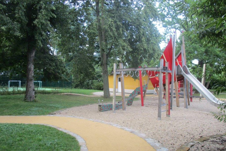 Zu sehen ist im Vordergrund ein großes Klettergerät mit mehreren Elementen zum Klettern und einer großen Rutsche. Im Hintergrund ist ein Sportplatz zu sehen.