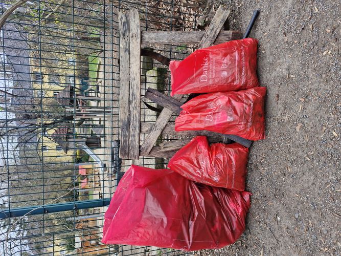 „Mehrere rote Müllsäcke stehen gefüllt vor einem Zaun neben alten Holzbrettern, im Hintergrund ein eingezäunter Spielplatz und Wohngebäude.“