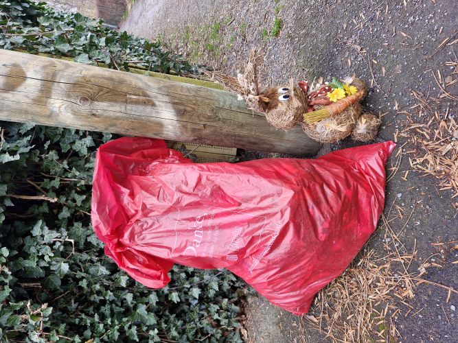 „Großer roter Müllsack lehnt an einem hölzernen Pfosten neben einem dekorativen Hasen aus Naturmaterialien, im Hintergrund ein mit Efeu bewachsener Zaun und ein befestigter Weg.“