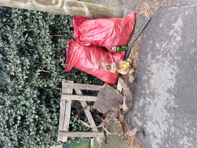 „Zwei große rote Müllsäcke sowie verschiedene aufgesammelte Metall- und Holzreste stehen vor einem mit Efeu bewachsenen Zaun; daneben ein dekorativer Hase aus Naturmaterialien und eine zurückgelassene Glasflasche.“