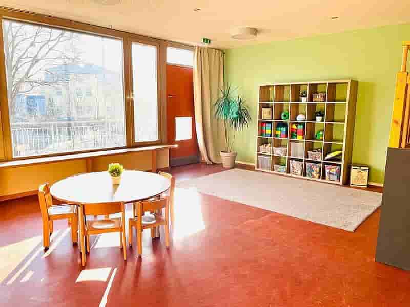 Entdeckerraum im Kindergarten – Kreativer Spiel- und Lernraum mit bunten Möbeln Heller Entdeckerraum im Kindergarten mit roten Boden, bunten Tischen und Stühlen, Spielmaterialien und großen Fenstern