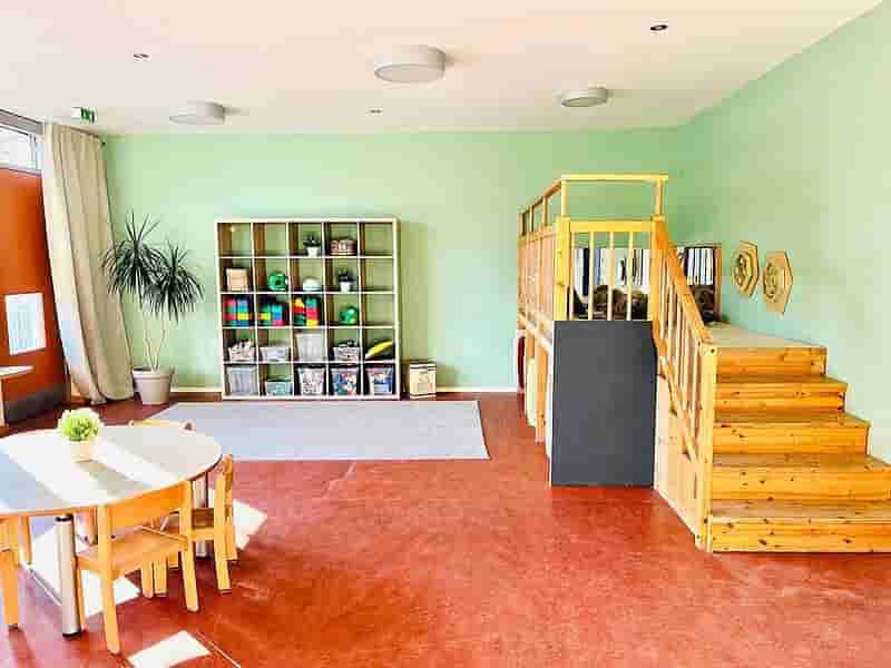 Entdeckerraum im Kindergarten – Klettertreppe, Spielbereich und kreative Lernumgebung Heller Entdeckerraum im Kindergarten mit rotem Boden, Holz-Klettertreppe, Regalen mit Spielmaterialien und Tischbereich für Kinder