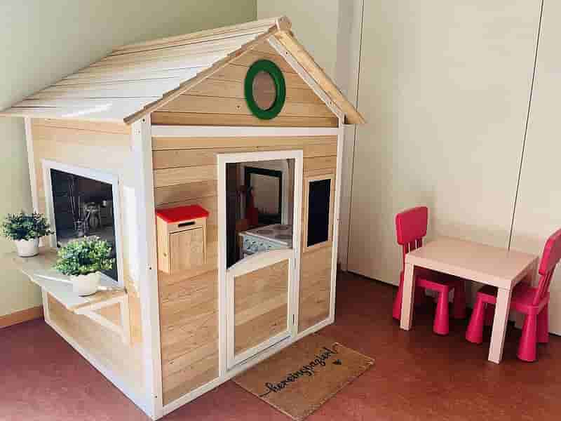 Spielhaus im Kindergarten – Rollenspielbereich mit Kaufladen und Kindertisch Holz-Spielhaus im Kindergarten mit Kaufladen-Fenstern, Spielküche im Inneren und pinkem Kindertisch daneben