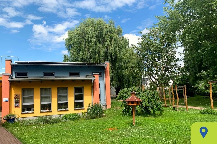 Einrichtung und Garten des Kindergarten Vieselbach in Erfurt Teil des Gebäudes und Eingangs des Kindergartens Vieselbach mit viel Grün und Bäumen, der Himmel ist blau mit einzelnen Wolken