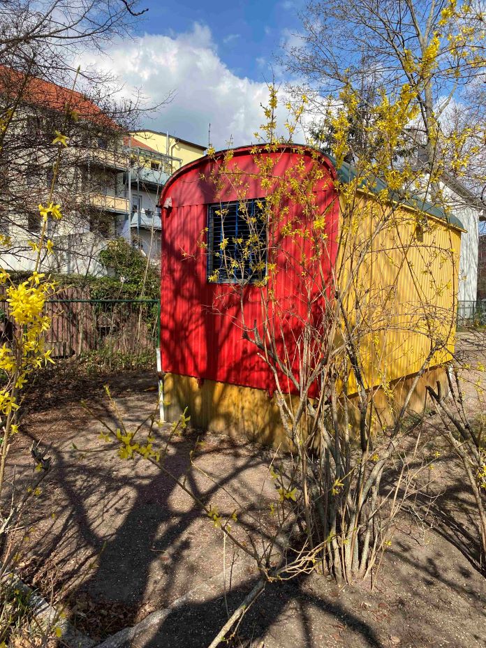 Roter und gelber Bauwagen im Außengelände des Kindergartens Vollbrachtfinken, umgeben von Sträuchern und Bäumen.