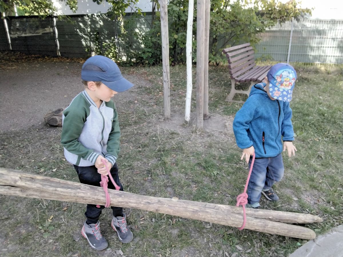 Naturmaterialien - Bauen mit Holz - Freispiel - Kindergarten Windischholzhausen Zwei Kinder tragen jeweils mit einem Seil zwei lange Holzpfähle