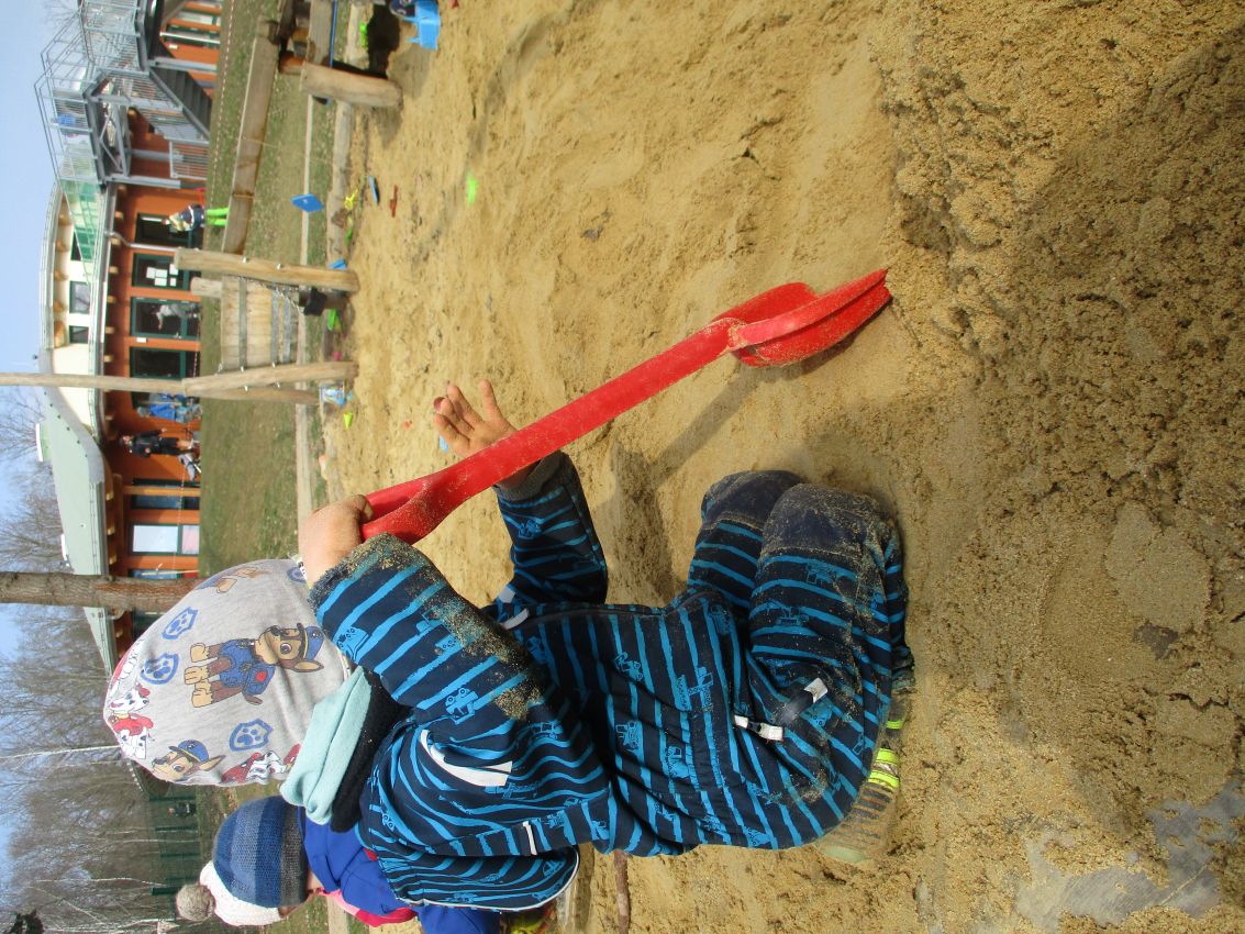  Ein Kind sitzt im Sandkasten. Es trägt einen blau gestreiften Anzug und eine bunte Mütze. Das Kind hält eine große rote Schaufel und gräbt im Sand. Im Hintergrund spielen andere Kinder. Dort steht auch ein Gebäude mit orange Wänden und grünem Dach. Es ist draußen, der Himmel ist grau.