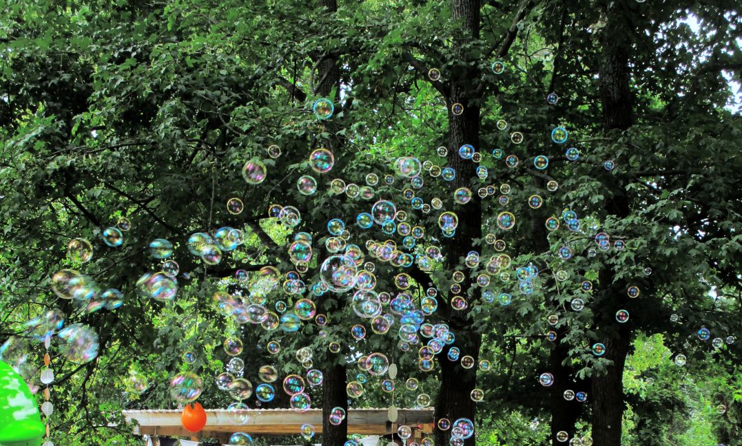 Gemeinsame Feste im Kindergarten Burgweg – Seifenblasen im grünen naturnahen Garten Viele bunte Seifenblasen schweben vor grünen Bäumen im naturnahen Garten des Kindergarten Burgweg. Im unteren Bildbereich sind Teile von Spielgeräten und Dekorationen sichtbar, die auf fröhliche Outdoor-Aktivitäten für die Kinder hinweisen.